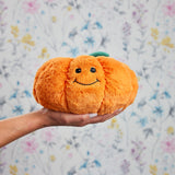 Une peluche bouillotte en forme de citrouille souriante, orange et douce, tenue dans une main devant un fond floral pastel.