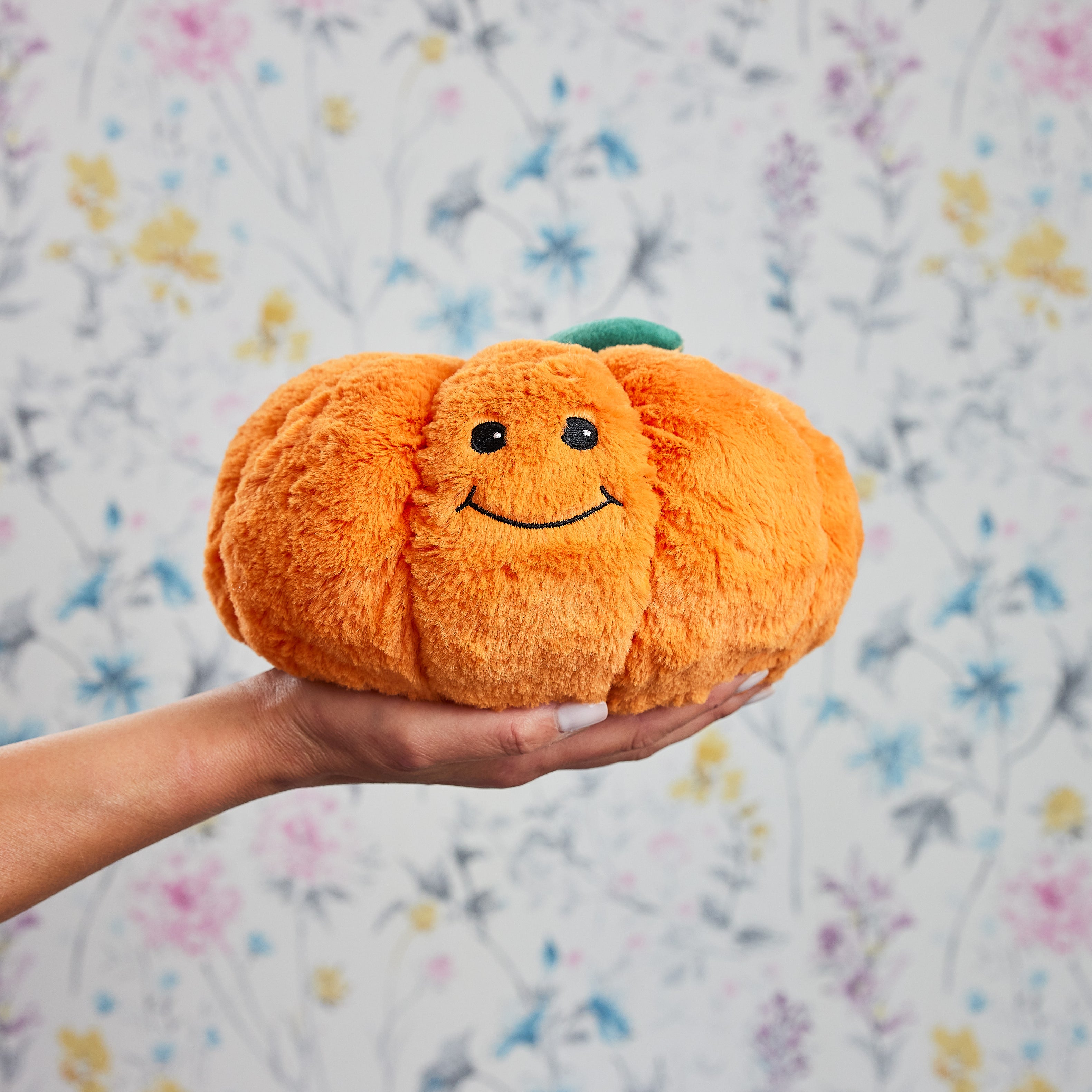 Une peluche bouillotte en forme de citrouille souriante, orange et douce, tenue dans une main devant un fond floral pastel.