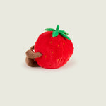 Bouillotte peluche en forme de fraise, rouge avec sur le haut deux feuille et une tige verte. Elle a un visage avec un sourire, des bras et des jambes marron. Elle est de profil 