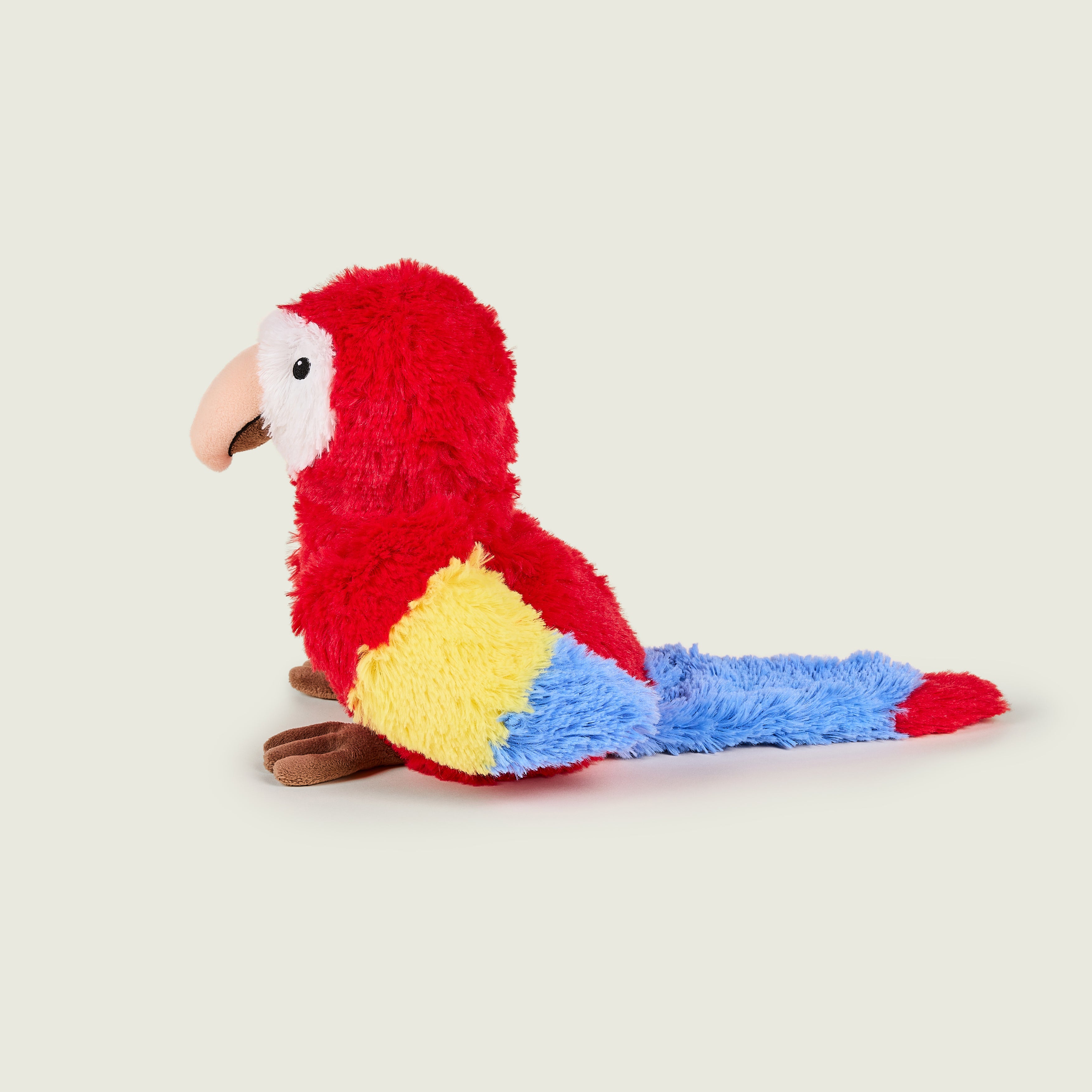 Bouillotte peluche en forme de perroquet, Pelage rouge et blanc sur la tête. Bec crème et pattes marrons. Bout des ailes jaune et bleu. il est de profil. 