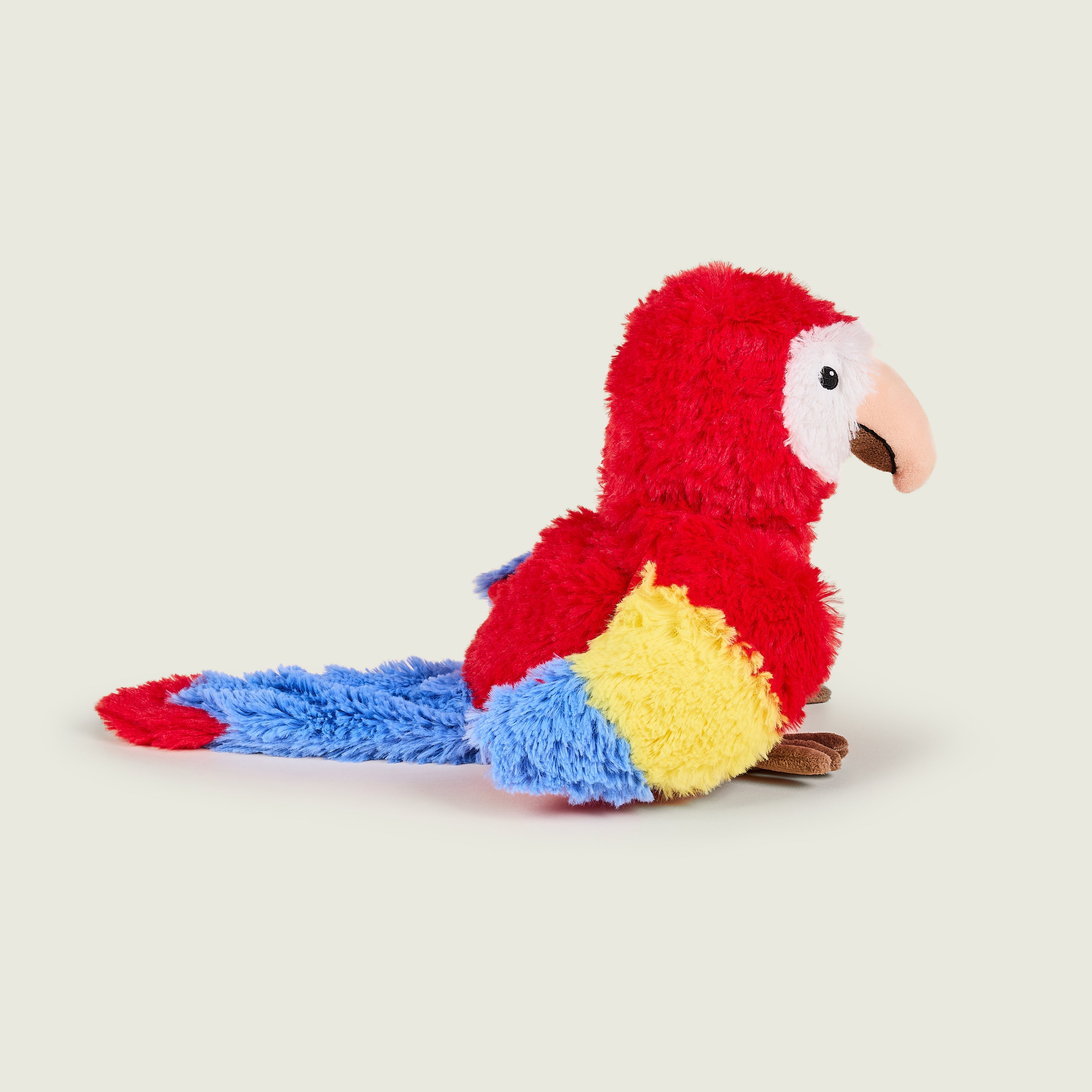 Bouillotte peluche en forme de perroquet, Pelage rouge et blanc sur la tête. Bec crème et pattes marrons. Bout des ailes jaune et bleu. Il est de profil. 