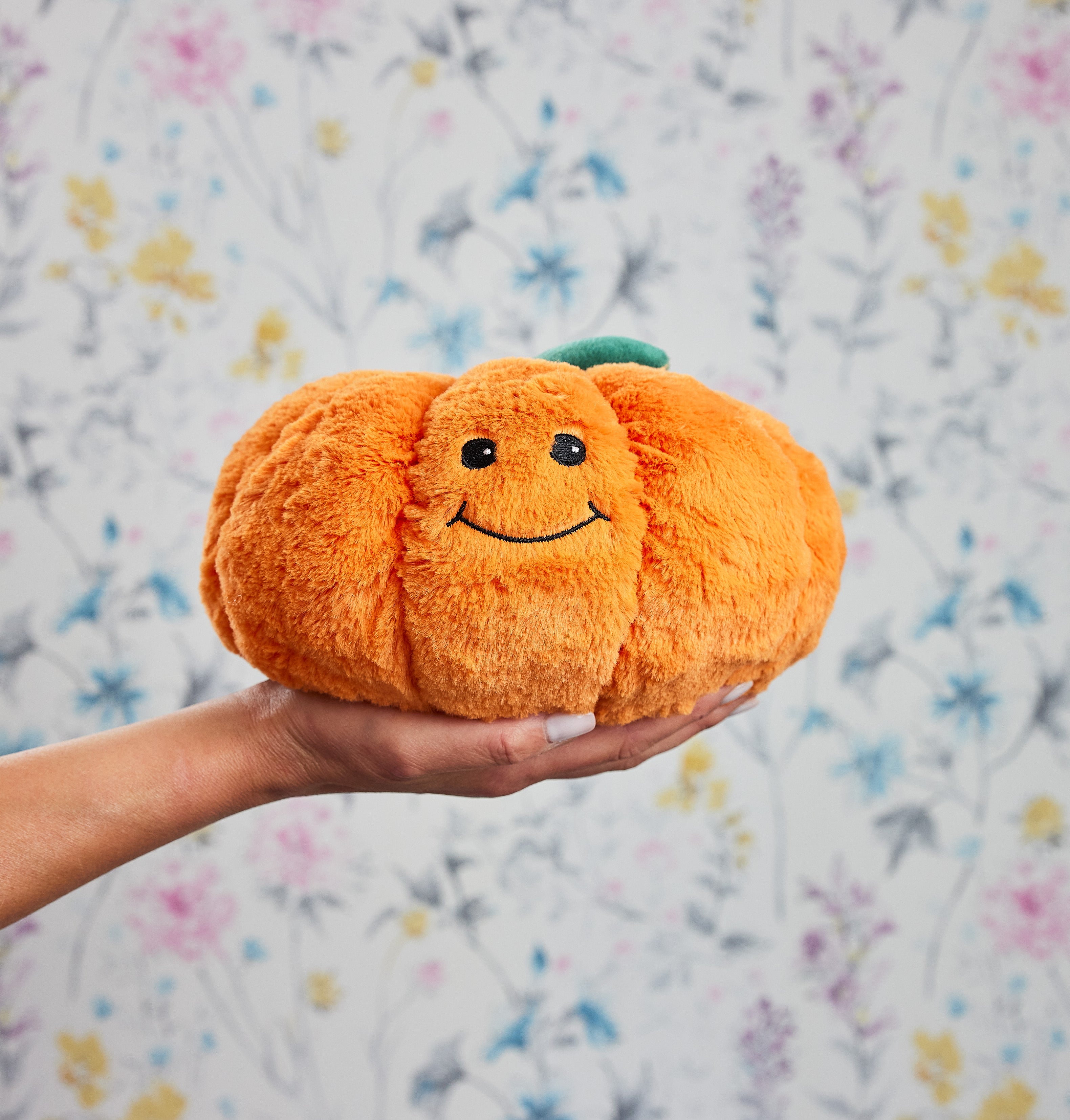 Une peluche bouillotte en forme de citrouille souriante, orange et douce, tenue dans une main devant un fond floral pastel.