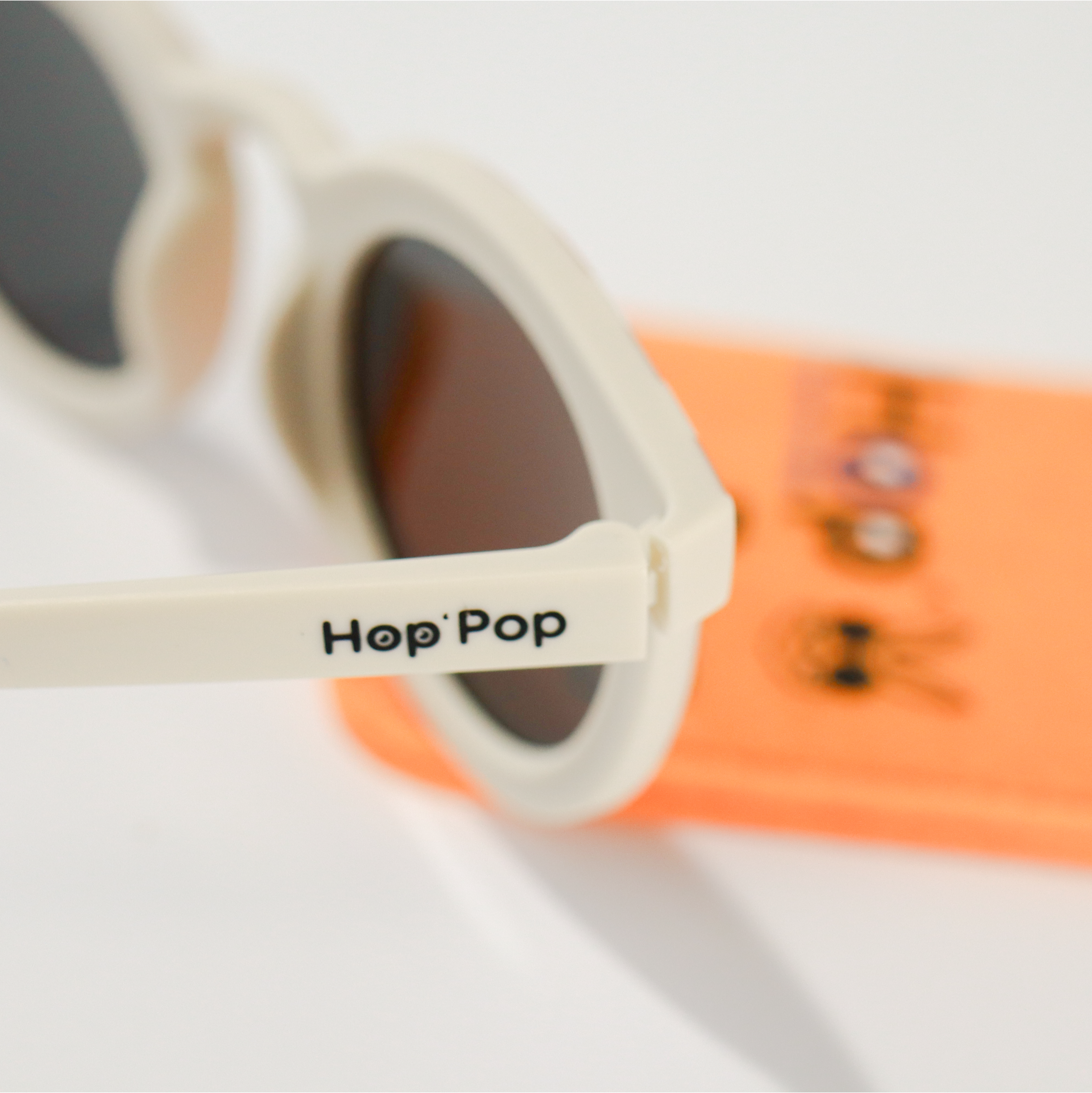 Gros plan sur une paire de lunettes de soleil crème de la marque Hop'Pop. Le logo de la marque est visible en noir sur la branche latérale. Les verres teintés, probablement polarisés, suggèrent une protection solaire élevée. À l’arrière-plan flou, on distingue une pochette de rangement orange vif avec des illustrations ludiques, assortie à l’univers enfantin ou familial de la marque.