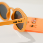 Gros plan sur une paire de lunettes de soleil orange de la marque Hop'Pop. Le logo de la marque est visible en noir sur la branche latérale. Les verres teintés, probablement polarisés, suggèrent une protection solaire élevée. À l’arrière-plan flou, on distingue une pochette de rangement orange vif avec des illustrations ludiques, assortie à l’univers enfantin ou familial de la marque.