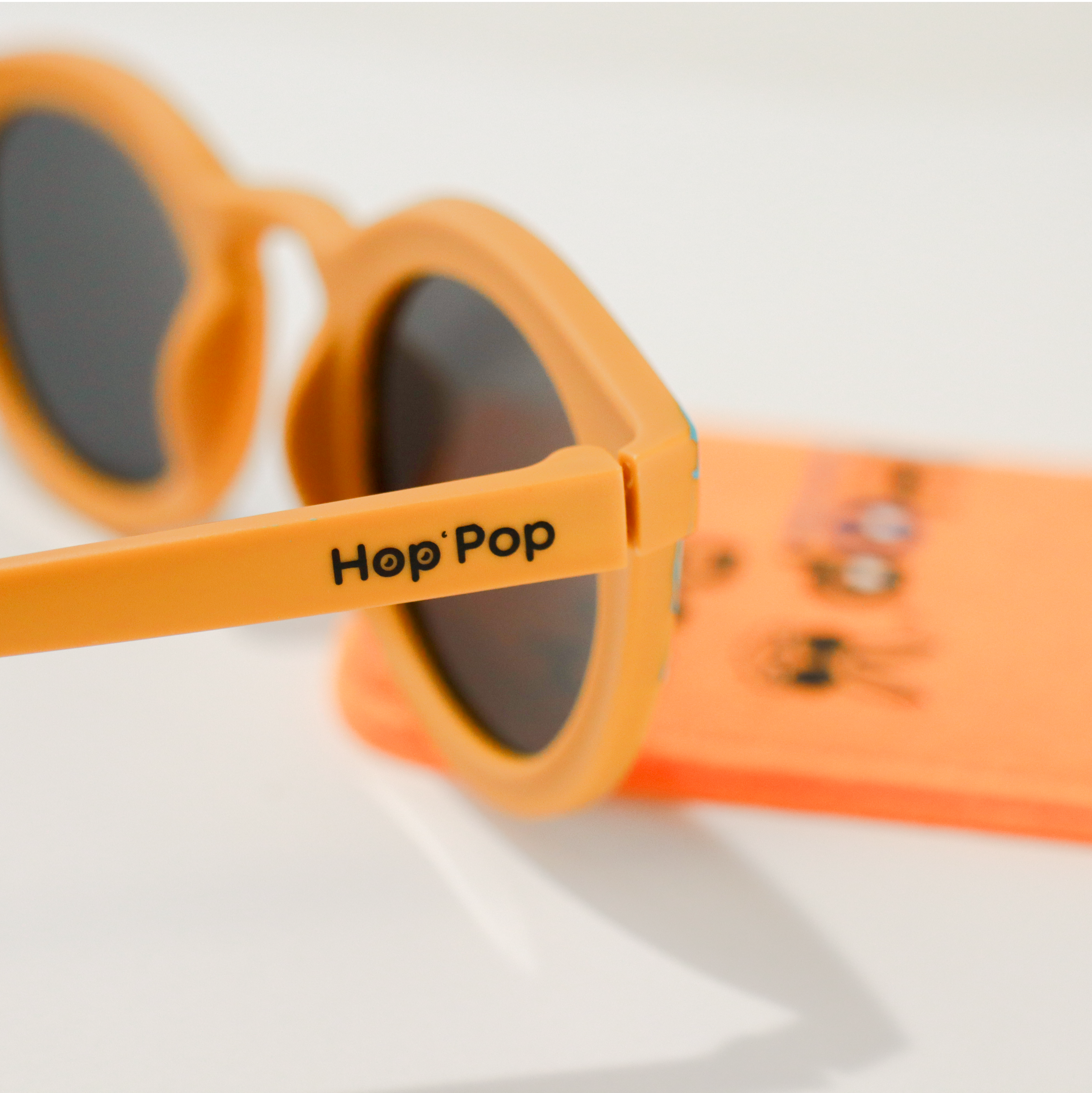 Gros plan sur une paire de lunettes de soleil orange de la marque Hop'Pop. Le logo de la marque est visible en noir sur la branche latérale. Les verres teintés, probablement polarisés, suggèrent une protection solaire élevée. À l’arrière-plan flou, on distingue une pochette de rangement orange vif avec des illustrations ludiques, assortie à l’univers enfantin ou familial de la marque.