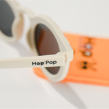 Gros plan sur une paire de lunettes de soleil crème de la marque Hop'Pop. Le logo de la marque est visible en noir sur la branche latérale. Les verres teintés, probablement polarisés, suggèrent une protection solaire élevée. À l’arrière-plan flou, on distingue une pochette de rangement orange vif avec des illustrations ludiques, assortie à l’univers enfantin ou familial de la marque.