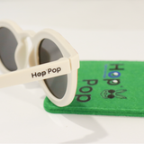 Gros plan sur une paire de lunettes de soleil crème de la marque Hop'Pop. Le logo de la marque est visible en noir sur la branche latérale. Les verres teintés, probablement polarisés, suggèrent une protection solaire élevée. À l’arrière-plan flou, on distingue une pochette de rangement verte avec des illustrations ludiques, assortie à l’univers enfantin ou familial de la marque.