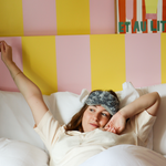 Jeune femme allongée dans un lit, portant un masque bouillotte en peluche gris Warmies sur le front, en train de s’étirer comme au réveil