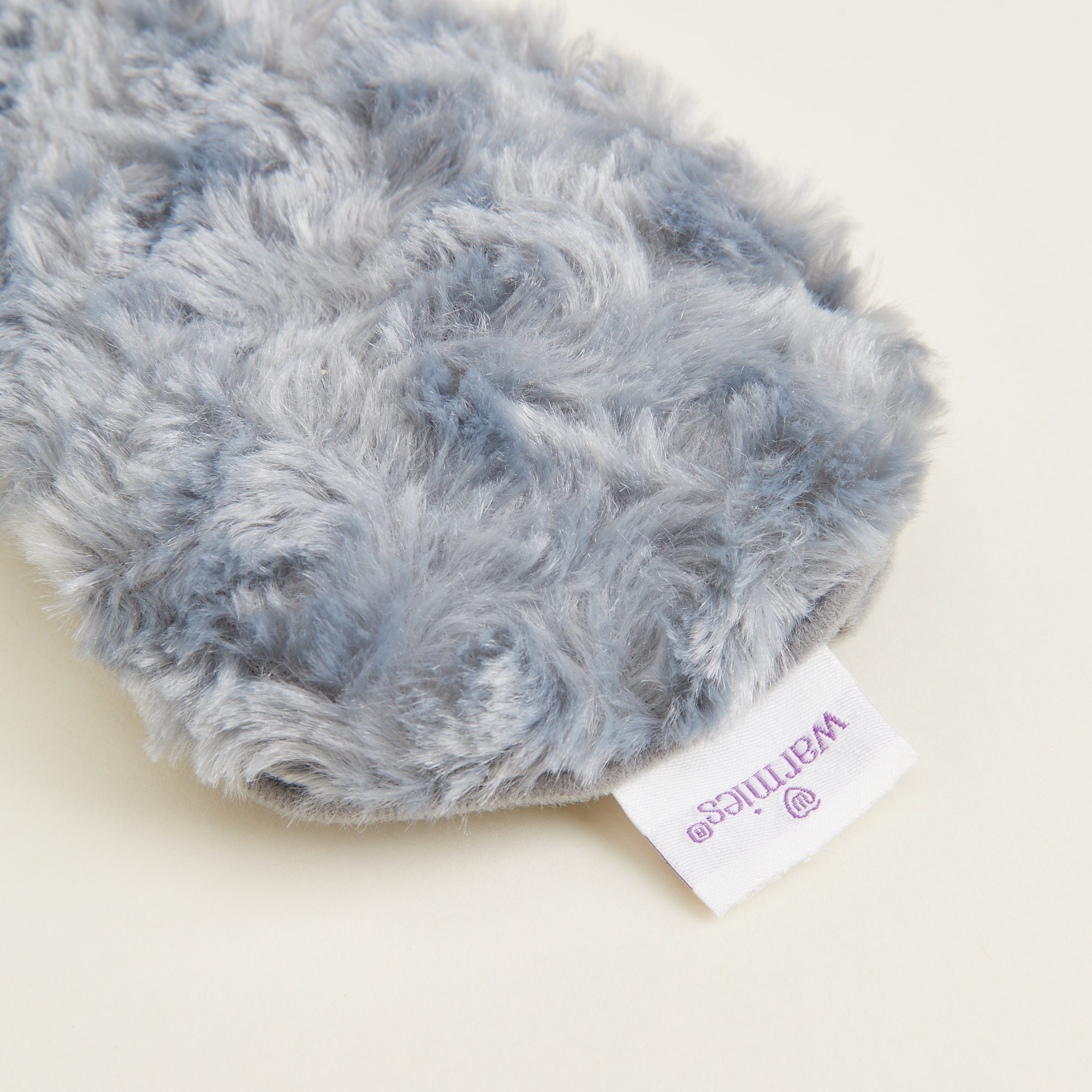 Gros plan sur la matière peluche bouclée du masque bouillotte  gris et l’étiquette Warmies