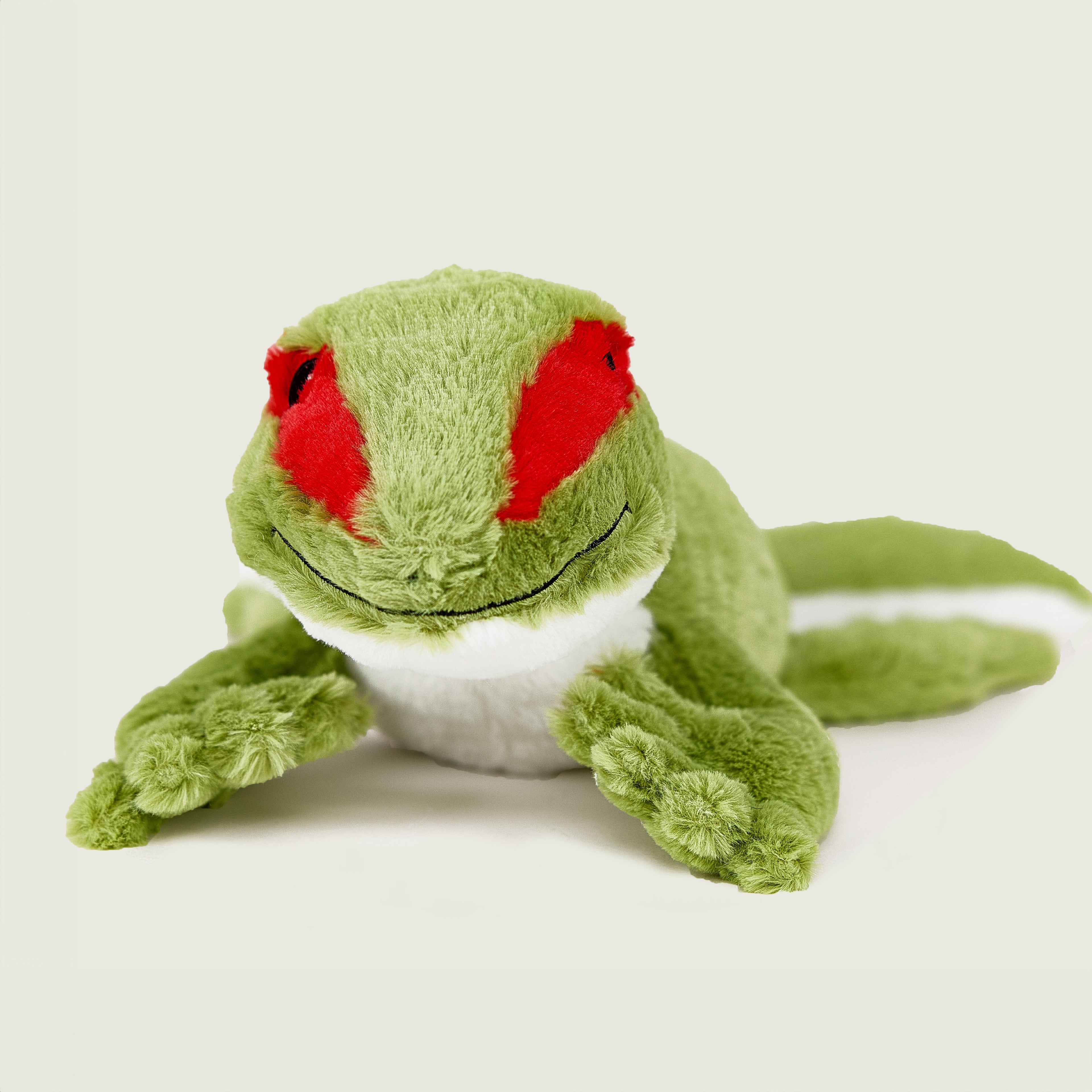 Peluche bouillotte déhoussable gecko verte, corps inférieur blanc, avec visage souriant et aplats rouge au niveau des yeux. Vue de face