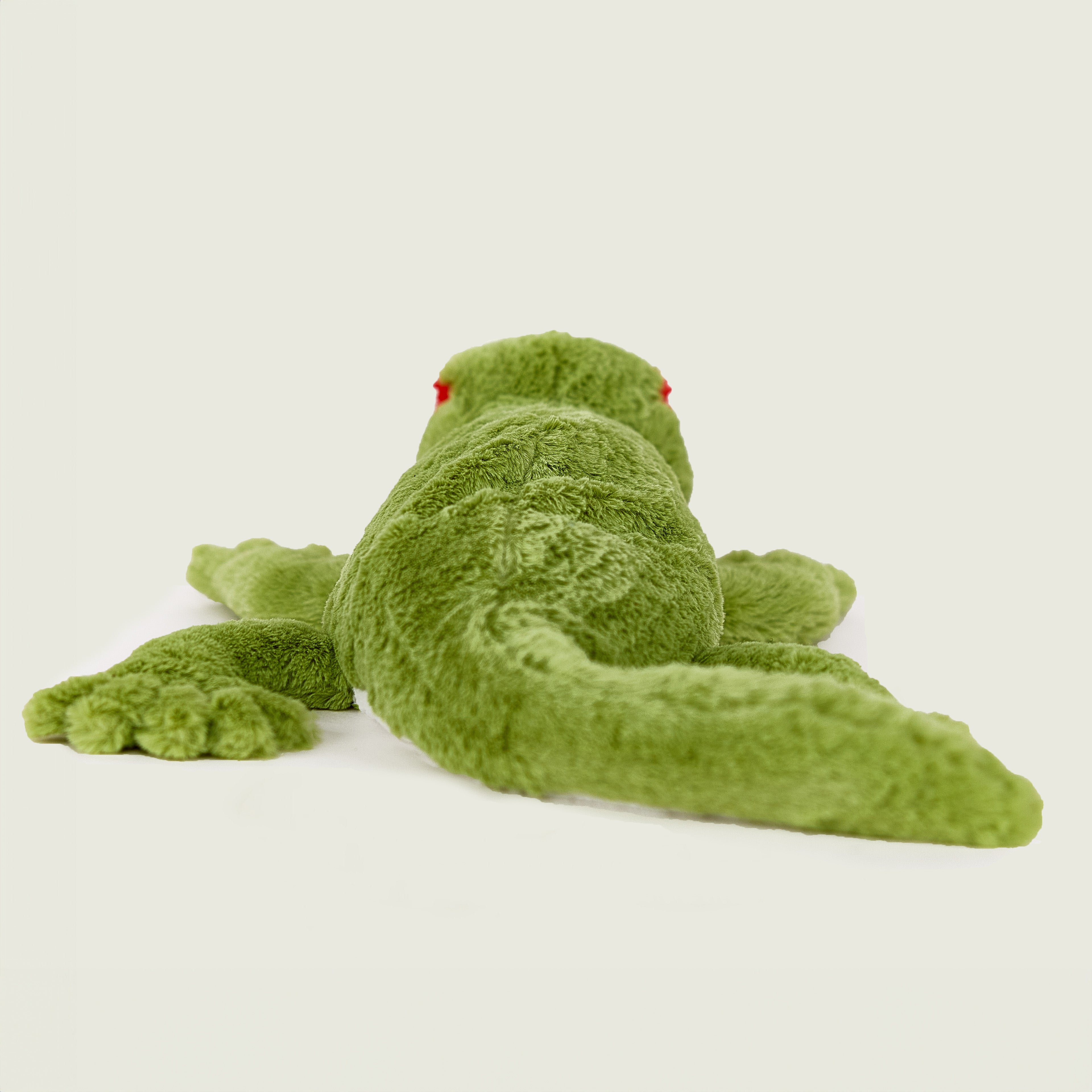 Peluche bouillotte déhoussable gecko verte, corps inférieur blanc, avec visage souriant et aplats rouge au niveau des yeux. Vue de dos et de la queue